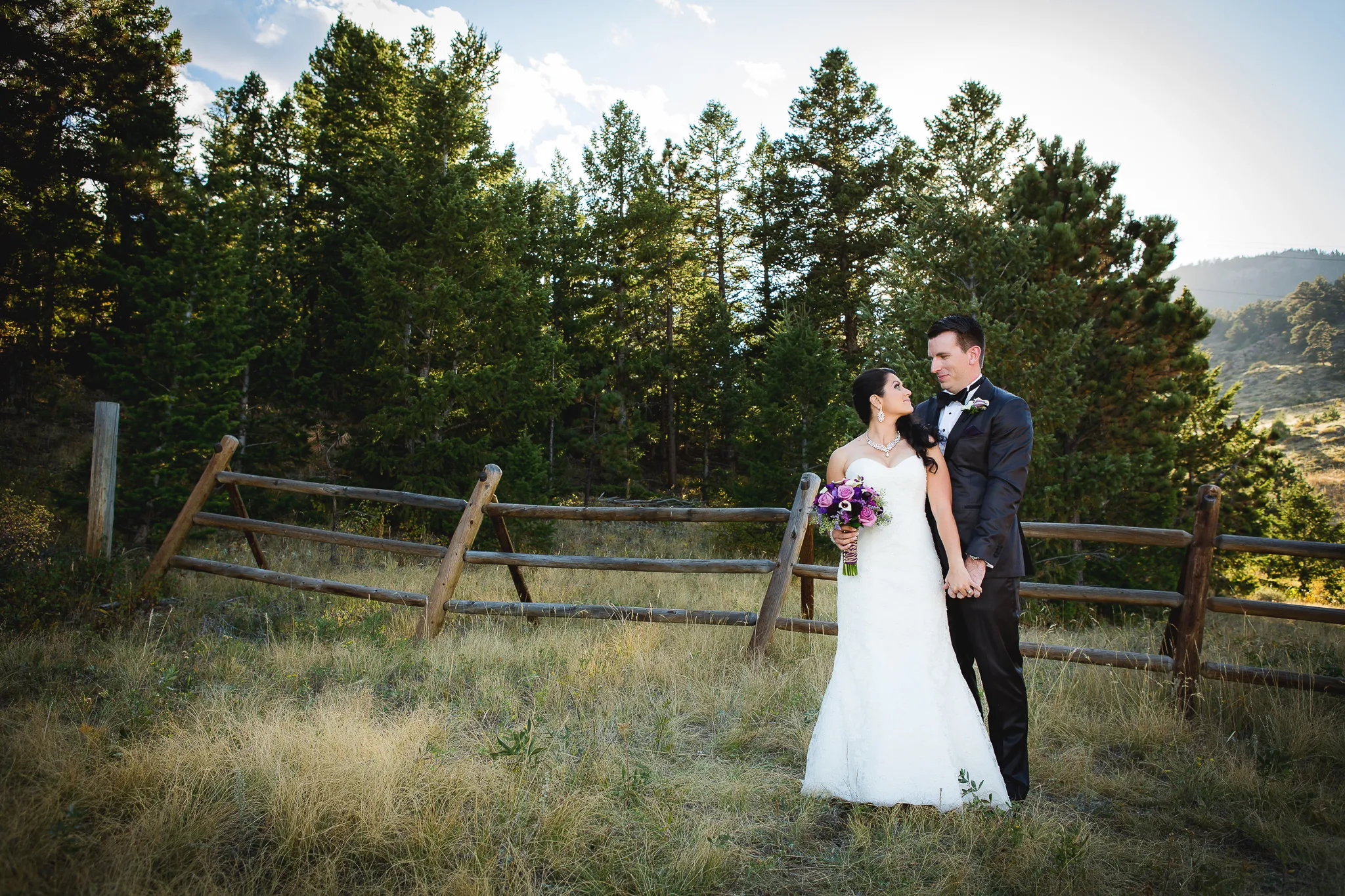 James and Samantha on their wedding day, in front of pines and a split-rail fence in the Colorado foothills